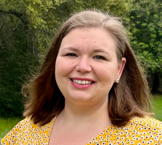 Headshot_McQueston: Dr. Jessica McQueston, Chair of the Special and Inclusive Education Research (SIER) SIG, stands outdoors in front of a backdrop of green trees and grass. She is a white woman in her late thirties. She is smiling warmly and wearing a yellow dress with a white and black floral pattern. Her shoulder-length brown hair is gently blowing in the breeze. The natural setting and her relaxed, confident expression create a welcoming and approachable tone. 
